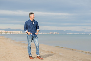 Handsome middle-aged man walking at the beach. Attractive happy smiling mid adult male model posing at seaside in blue jeans, t-shirt shirt. Full-length outdoor portrait of beautiful macho man.