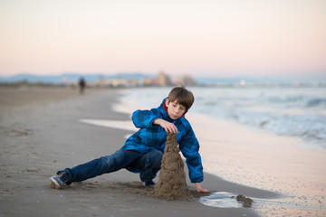Fototapeta na wymiar Young boy builds sand castle at the winter beach. Cute 11 years old boy at seaside, evening time. Kid's outdoor portrait.