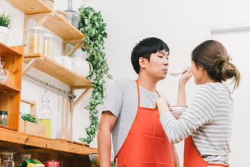 Young Asian lovely couple cook together at home kitchen, tasting food preparing meal. Girl feed soup to boyfriend using spoon. Married people lifestyle, love relationship or domestic life concept