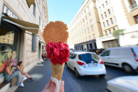 Appetizing pink berry ice cream in waffle cone in female hand over Rome city view. Tasty ice cream like a symbol of summer and italian dessert over roman street.