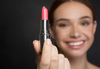 Close up of pink lipstick in hand of positive girl who is demonstrating it to the camera. Female with smile is on background. Isolated and selective focus. Beauty concept