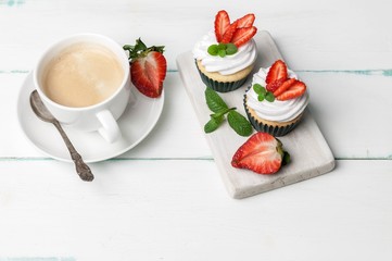 Vanilla cupcakes with butter cream and fresh strawberry and a cup of coffee on a light wooden background