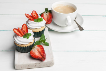 Vanilla cupcakes with butter cream and fresh strawberry and a cup of coffee on a light wooden background