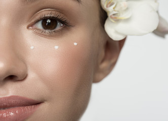 Healthy skin concept. Close up half of face of positive attractive young woman with orchid is looking at camera with slight smile. Focus on eye with drops of moisturizer emulsion are around