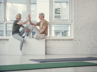 Side view profile of aging woman and gentleman sitting opposite each other on windowsill, they are greeting each other with a high five and laughing. Copy space in right side