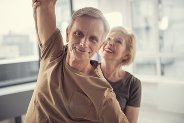 Portrait of glad slim pensioner with raised arm. Smiling senior woman on background holding his arm. Focus on man