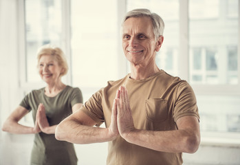 Waist up portrait of joyful old male standing with clasped hands. Woman in same posture on background. Focus on man