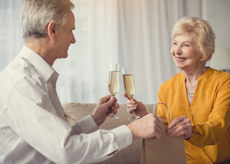 Smiling mature man and woman resting on sofa with alcohol in glasses. Male passing package with present to female
