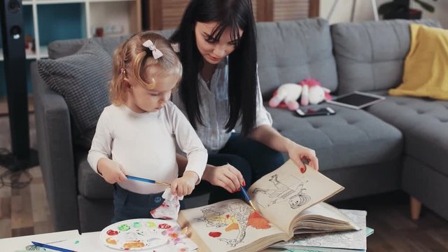mother with daugther painting on paper book hold paintbrush hug sit on sofa together at home happy kid family hobby girl love house coloring mom play art childhood table artist closeup cute