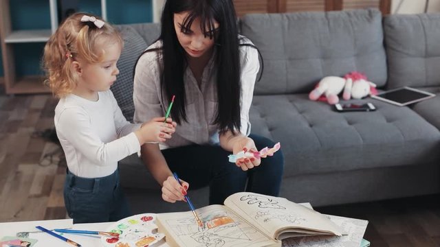 Portrait beautiful mother with daugther painting on paper book hug sit on sofa together at home happy kid family hobby girl love house coloring mom play art childhood table artist closeup slow motion