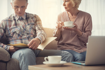 Tranquil senior man holding and reading documents, wife sitting next to him with cup and listening with attentiveness