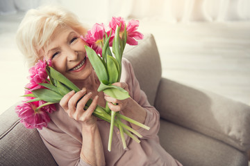 Elderly female person sitting at home with tulips and looking at camera with bliss. Copy space in right side. Top view