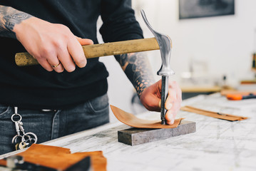 Close up of a leather craftsman working with leather using hammer. Working process in the leather workshop. Man holding crafting tool and working.
