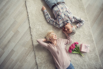 Top view of delighted old man and woman reposing on mild carpet and looking at each other with fondness. Flowers and present lying next to them. Copy space in left side