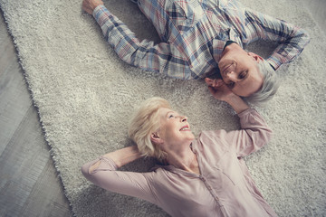 Top view of satisfied senior couple resting on comfortable rug and looking at each other with...