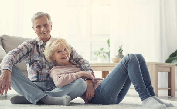 Sweet home. Portrait of cheerful senior couple resting on soft carpet in living room. They are looking at camera and laughing