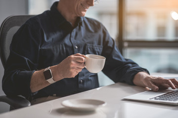 Smiling retire male worker tasting cup of coffee while sitting at table. He working with laptop