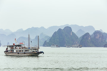 Cruising tourist boat over emerald water with towering limestone islands in the background in summer at Quang Ninh, Vietnam.