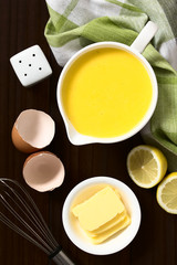 Hollandaise sauce, a basic sauce of the French cuisine, served in a sauce boat with ingredients (egg, butter, lemon) and whisk on the side, photographed overhead on dark wood with natural light