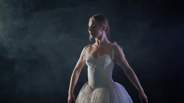 graceful ballerina in white tutu dress dancing elements of classical or modern ballet in the dark with light and smoke on the black background, slow motion