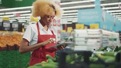 Serious african american woman seller uses the tablet to work in the vegetable department light at supermarket black industry worker grocery market healthy staff produce job service retail store shop - Powered by Adobe