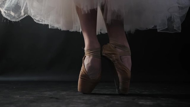 Close up of ballet dancer as she practices exercises on dark stage or studio. Woman's feet in pointe shoes. Ballerina shows classic ballet pas. Slow motion. Flare, gimbal shot.