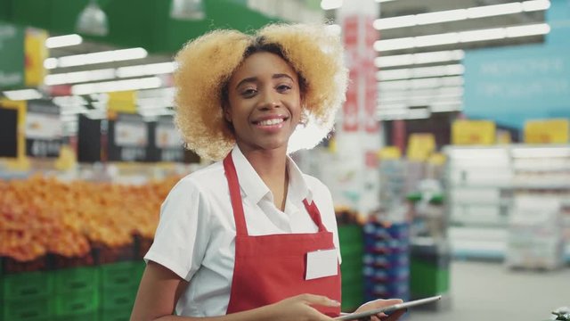 Smiling african american woman seller uses tablet to work in the vegetable department look at camera light at supermarket black industry worker grocery market staff job retail store shop