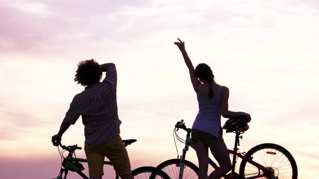 Young couple with bicycles at sunset. Young man and woman waving with hands at sunset, back view. Enjoying the horizon.
