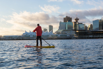 Adventurous man is paddle boarding near Downtown City during a vibrant winter sunrise. Taken in...