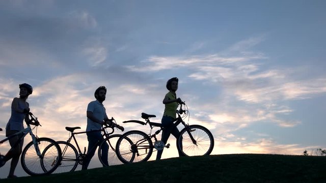 Young cyclists walking on hill at sunset. Group of friends with bikes on beautiful sky background. Amazing rest outdoors.