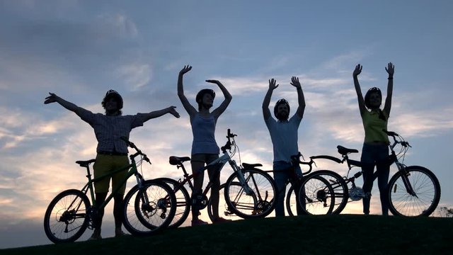 Company of young cyclists at sunset sky. Group of young people with bicycles waving with hands on sky background. Best rest outdoors.