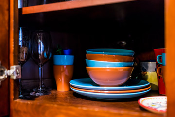 Open wooden kitchen cabinet door cupboard with many colorful dishes, bowls, plates, cups, glasses on shelves closeup