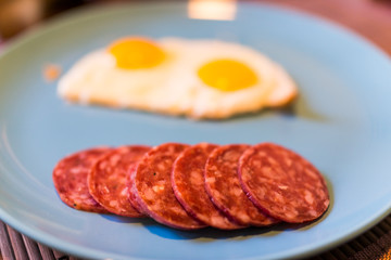 Closeup of two fried eggs on large blue plate with cured meat hard salami slices
