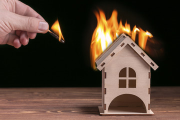 Model house in fire and hand with matchstick on a black background.