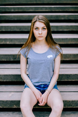 Fototapeta premium Young girl portrait. Blue eyed brunette female expressive sensual face. Big sad innocent eyes of adorable woman sitting on wooden stairs outdoor. Reflexive person state of mind. Deep inner mood.