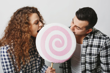 Happy female and male have fun together, bite big round lollipop, likes sweets, isolated over white background. Couple in love enjoy eatting delcious candy. Togetherness and relationship concept