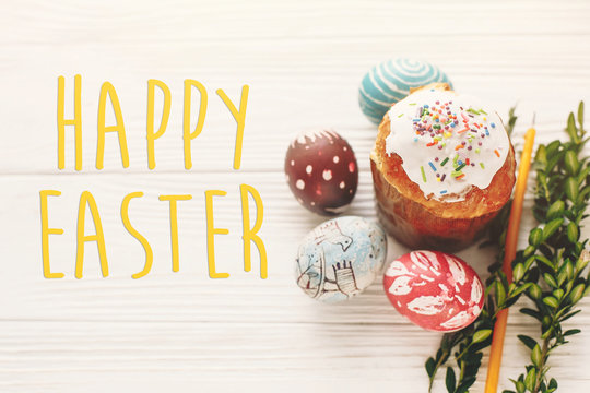 happy easter text. season's greetings card. stylish painted eggs and easter cake on white rustic wooden background with spring flowers and candle. modern easter image
