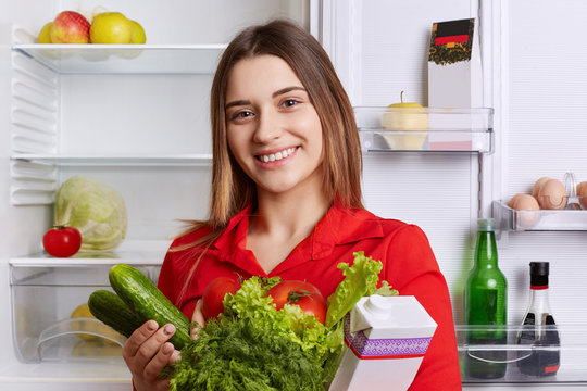 Satisfied woman holds fresh vegetables and milk, going to put them in refridgerator, has broad smile and happy expression, being vegeterian, eats only healthy food. People and nutrition concept