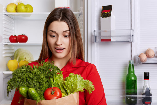 Amazed young female with surprised expression looks at vegetables, forgets to buy something in grocer`s shop, stands in kitchen near refrigerator. People, healthy nutrition and dieting concept.
