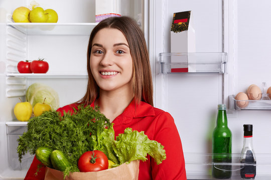 Pretty young female with cheerful expression, has broad smile, holds paper bag full of fresh vegetables: red tomatoes, cucumbers, dill, lettuce, stands in kitcen. Cooking and dieting concept