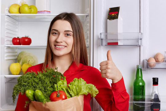 Pleasant looking of young woman with long straight hair, dressed in red blouse, holds paper bag with vegetables, shows ok sign, stands near fridge, has delighted look. People, nutrition concept