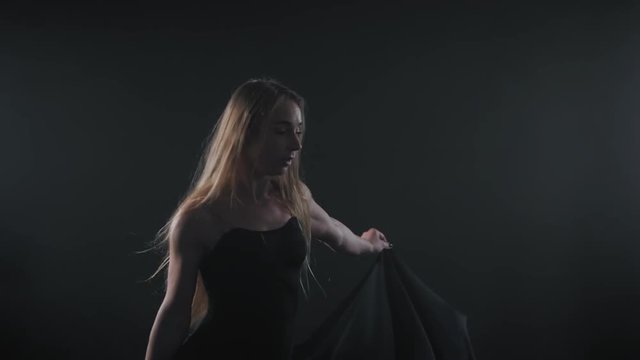 Professional ballerina dancing ballet in spotlights smoke on big stage.Beautiful caucasian young girl with long hair wearing black tight dress on floodlights background. slow motion