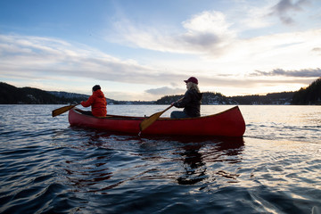 Fototapeta premium Couple friends on a wooden canoe are paddling in an inlet surrounded by Canadian mountains during a vibrant sunset. Taken in Indian Arm, near Deep Cove, North Vancouver, British Columbia, Canada.