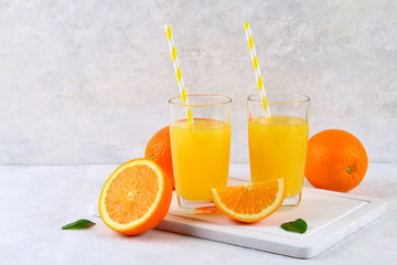 Glass cups and a pitcher of fresh orange juice with slices of orange and yellow tubes on a light gray table.