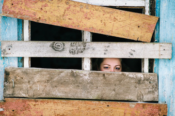 Locked up illegal refugee girl in abroad shelter portrait. Woman slavery. Werid strange unusual person looking through slit in boarded up window in old wooden abandoned ghost house. Social problems.