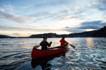 Couple friends on a wooden canoe are paddling in an inlet surrounded by Canadian mountains during a vibrant sunset. Taken in Indian Arm, near Deep Cove, North Vancouver, British Columbia, Canada.