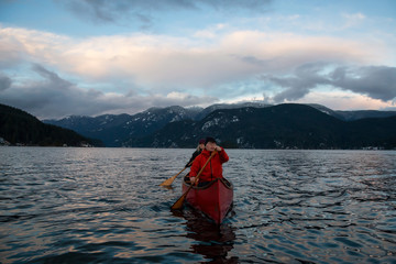 Naklejka premium Couple friends on a wooden canoe are paddling in an inlet surrounded by Canadian mountains during a vibrant sunset. Taken in Indian Arm, near Deep Cove, North Vancouver, British Columbia, Canada.