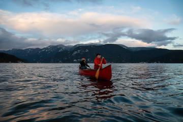 Couple friends on a wooden canoe are paddling in an inlet surrounded by Canadian mountains during a vibrant sunset. Taken in Indian Arm, near Deep Cove, North Vancouver, British Columbia, Canada.