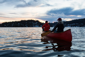 Couple friends on a wooden canoe are paddling in an inlet surrounded by Canadian mountains during a vibrant sunset. Taken in Indian Arm, near Deep Cove, North Vancouver, British Columbia, Canada.