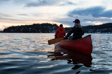 Couple friends on a wooden canoe are paddling in an inlet surrounded by Canadian mountains during a vibrant sunset. Taken in Indian Arm, near Deep Cove, North Vancouver, British Columbia, Canada.